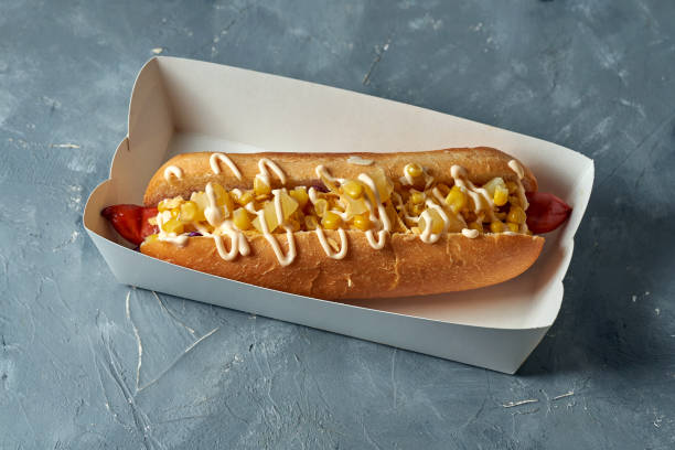 istock Appetizing hot dog with corn, pineapple, smoked sausage in a white box on a gray background 1319914475