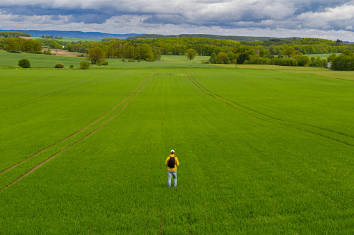 Aeirial view young caucasian man from back with backpack walking on spring green agriculture field. Czech landscape