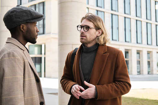 Young businessman in coat and in eyeglasses talking to his colleague on the street