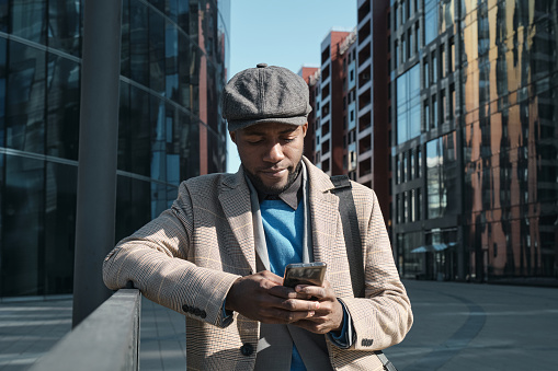 African businessman in stylish clothes communicating online on mobile phone while standing in the city