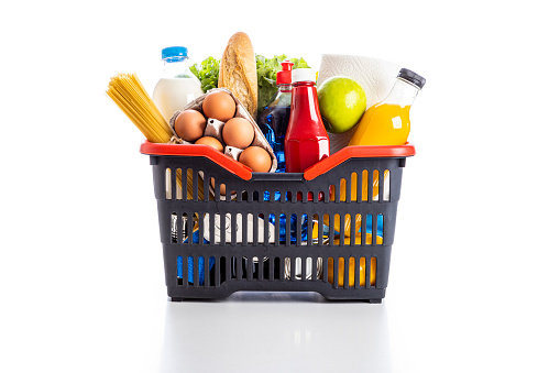 Front view of a black plastic shopping basket full of groceries such as a milk bottle, a ketchup bottle, an orange juice bottle, some eggs, pasta, a lettuce and a bread. The shopping basket is isolated on white background.
