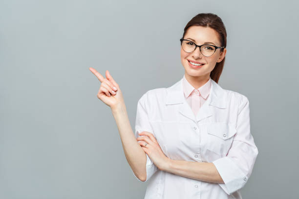 Friendly smiling young female doctor. Hand pointing at copy space. Isolated on a grey background Friendly smiling young female doctor. Hand pointing at copy space. Isolated on a grey background pharmacist stock pictures, royalty-free photos & images