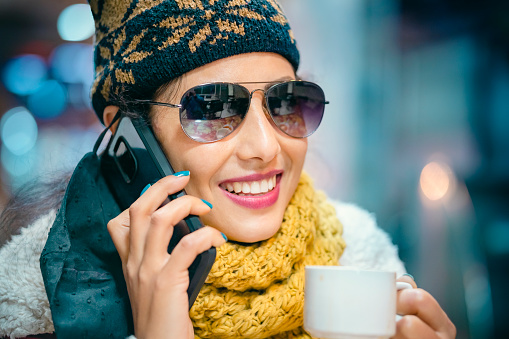 In this close-up indoor image with copy space, an Asian/Indian young woman talks on a smartphone while enjoying a cup of coffee in a restaurant at the time of the pandemic. She wears a face mask hanging on one ear, sunglasses, and warm clothing.