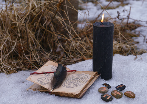Magic ritual with black candle, runes on stones and open with book with spells on snow. Esoteric, gothic and occult background, Halloween mystic and wicca concept outdoors.