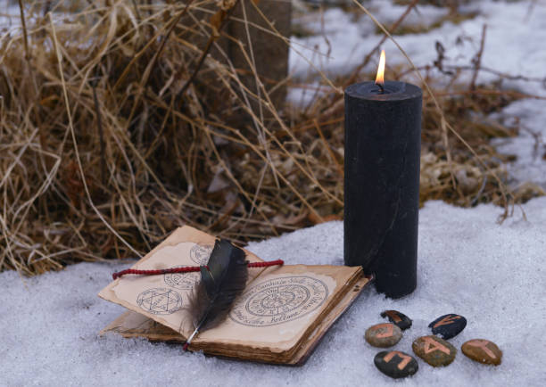 Magic ritual with black candle, runes on stones and open with book with spells on snow. stock photo