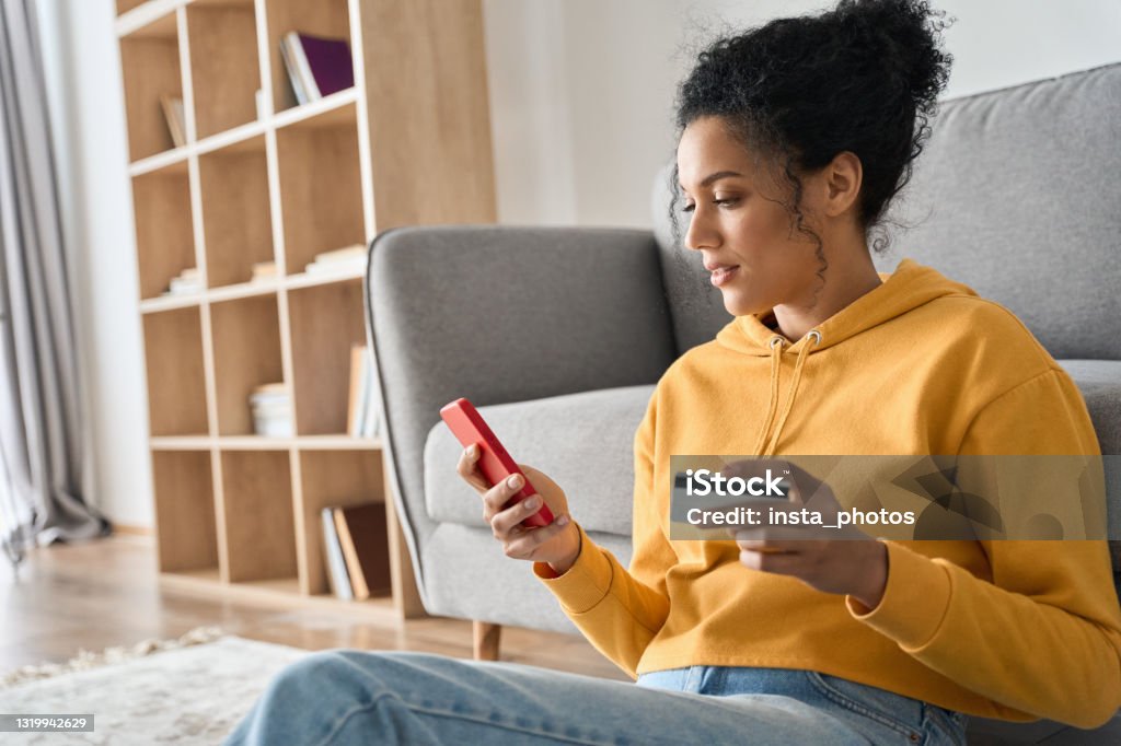 Young adult African american girl sitting indoors doing mobile payment online. Young adult African American female consumer holding credit card and smartphone sitting on floor at home doing online banking transaction. E commerce virtual shopping, secure mobile banking concept. Credit Card Stock Photo