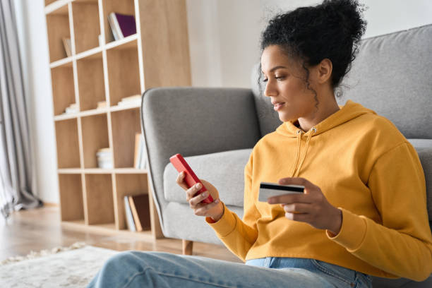 Young adult African american girl sitting indoors doing mobile payment online. Young adult African American female consumer holding credit card and smartphone sitting on floor at home doing online banking transaction. E commerce virtual shopping, secure mobile banking concept. credit card stock pictures, royalty-free photos & images