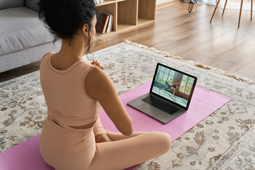 Young healthy African American mixed race girl wears sportswear meditate namaste hands watching online yoga class tutorial on pc at home sit on mat doing fitness exercise feeling no stress harmony.