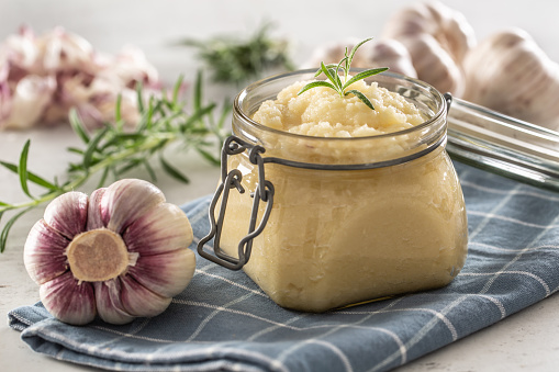 Aromatic garlic paste in a glass jar laid on rustic kitchen cloth with bulbs and peeled cloves and rosemary.