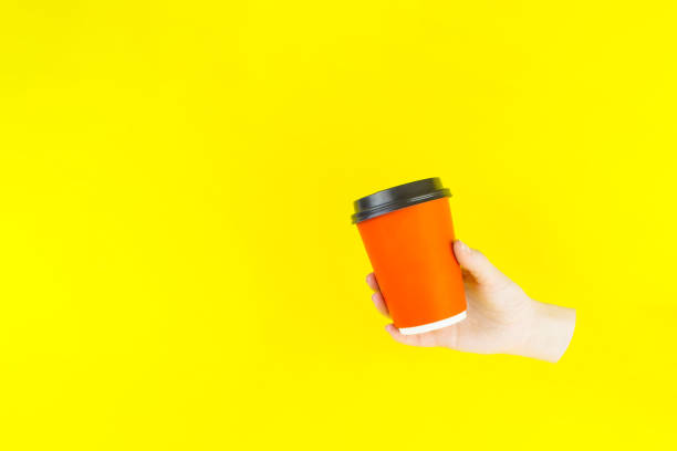 istock Hand Holding Orange Paper Cup on yellow background. Craft paper coffee cup with copy space 1319951995