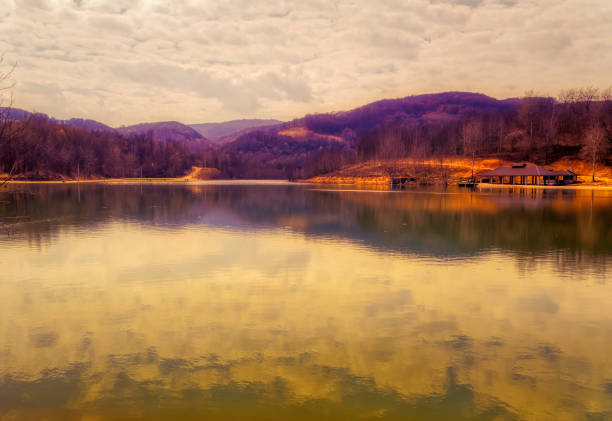 View over calm water of mountain lake surrounded by forest during cloudy spring day.