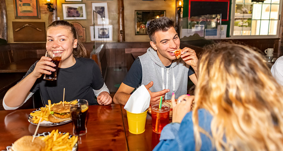Group of millennials friends eating at fast food indoors in irish pub restaurant. Happy people partying and eating together. Young active teenagers with burgers, pizza and drinks having fun dining.