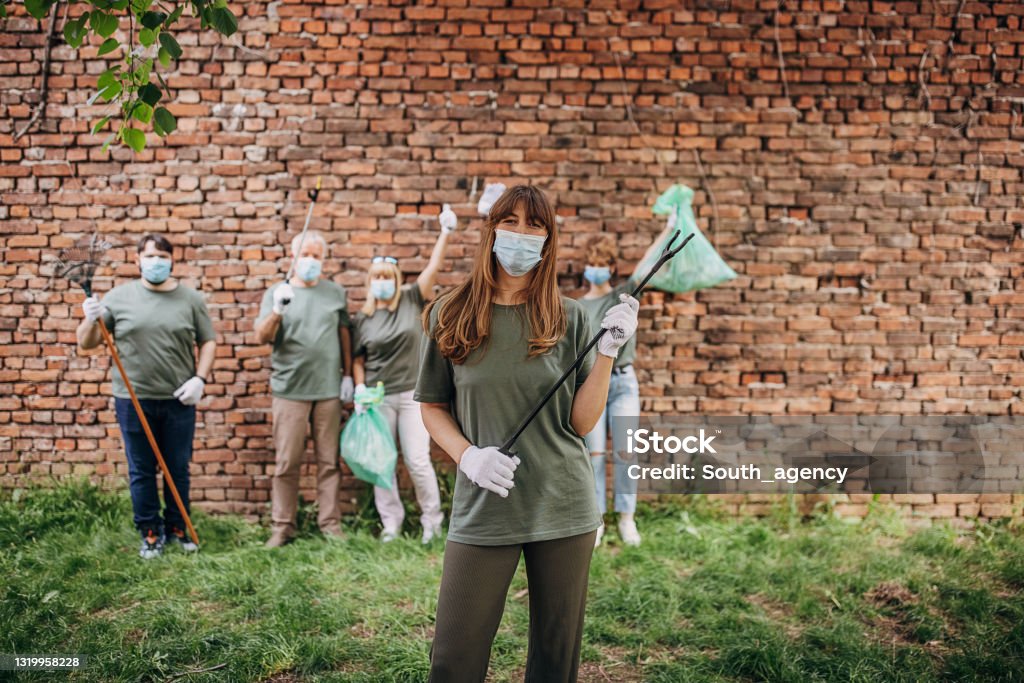 Group of volunteers Group of people, volunteers with garbage bags standing in a row by the brick outdoors, one woman is standing on front of them. They are all wearing protective face masks. Grabber Tool Stock Photo