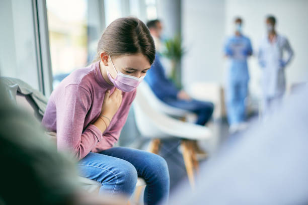 Sick little girl with face mask coughing while waiting for medical exam at the hospital. Small girl feeling ill and holding her chest in pain while sitting in waiting room at medical clinic during coronavirus pandemic. chest pain stock pictures, royalty-free photos & images