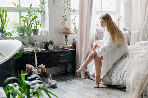 Beauty concept. Smiling young woman in bathrobe sitting on bed, apply body lotion or cream on skin at soft shaved legs. Female perform daily skincare routine spending morning in bedroom