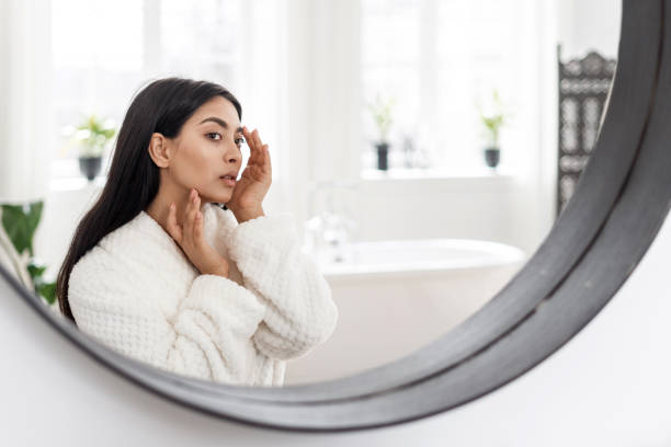 Young asian woman looking at mirror, checking skin Skincare and beauty concept. Side portrait view of young asian woman touching healthy facial skin, checking acne. Female in white bathrobe looking at mirror, spending morning in bathroom looking in mirror stock pictures, royalty-free photos & images