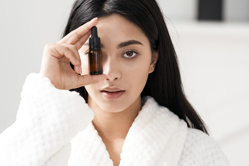 Portrait of young attractive asian woman holding skincare product in glass bottle with pipette in front of eye. Female advertising spa treatment as part of beauty routine. Organic cosmetic concept