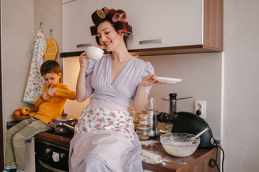Mother with the big hair curlers on her head sitting on the kitchen counter taking a coffee break while cooking with her son sitting on the kitchen counter licking a lollipop