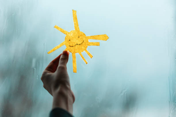 la mano di un bambino tiene il sole, tagliato fuori dal feltro, contro la finestra. fuori dalla finestra, pioggia e maltempo. primo piano. il concetto di depressione e buon umore. - drop window water fog foto e immagini stock