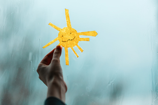 A child's hand holds the sun, cutted out of felt, against the window. Outside the window, rain and bad weather. Close-up. The concept of depression and good mood