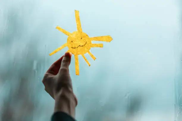 Photo of A child's hand holds the sun, cutted out of felt, against the window. Outside the window, rain and bad weather. Close-up. The concept of depression and good mood.