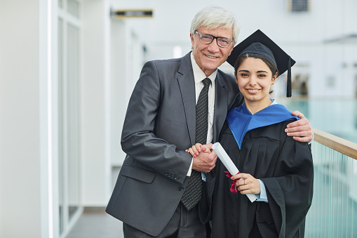 Waist up portrait of smiling young woman posing with father during graduation ceremony indoors