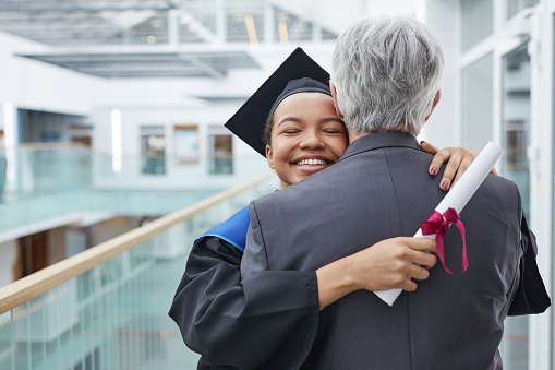 Portrait of happy African-American woman wearing graduation gown hugging father or college professor indoors, copy space