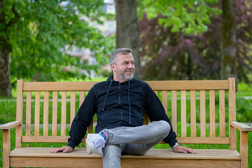 Man enjoying some quality time alone relaxing on a park bench in jeans and hoodie looking away to the side with a smile of pleasure