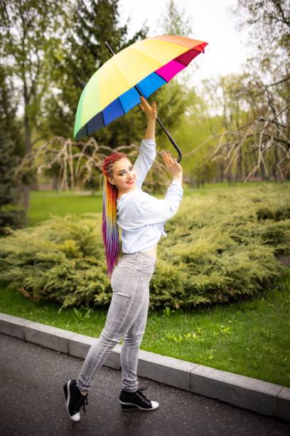istock A cute girl with multi-colored braids and bright makeup in a bluish shirt posing with a rainbow umbrella against the background of a blooming spring park 1319968536