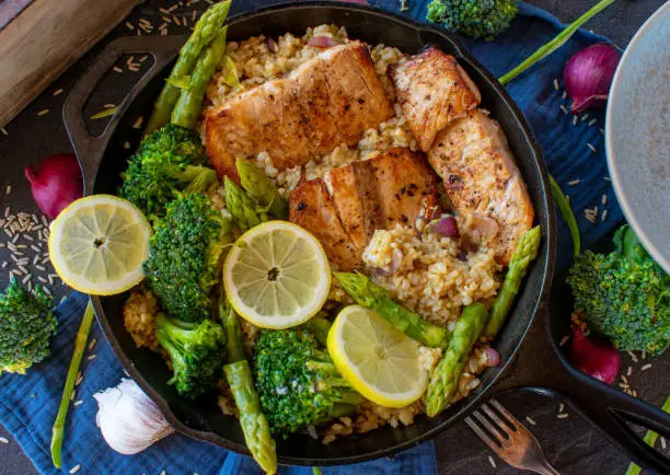 grilled salmon fillet with brown rice and green vegetables such as broccoli and asparagus served in a rustic cast iron pan on dark table background from above
