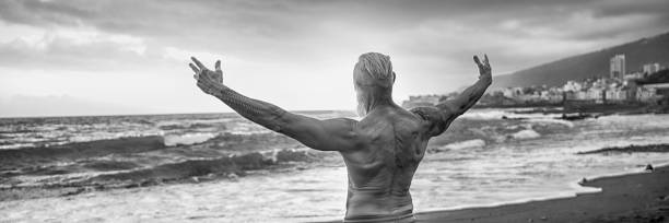 Age is just a number. In a healthy body, healthy mind. Senior man showing his muscular fit body Age is just a number. In a healthy body, healthy mind. Senior man showing his muscular fit body with tattoos on the beach. Summer vacation workout. Elderly people healthy lifestyle on a retirement concept. arm tattoo ideas for men pictures stock pictures, royalty-free photos & images