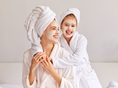 Cheerful girl in bathrobe and towel smiling and embracing young woman with moisturizing mask while resting on bed at home