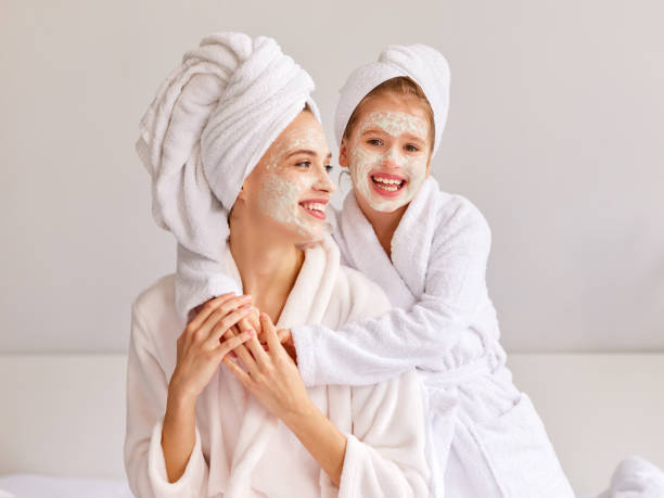 Happy mother and daughter with moisturizing mask Cheerful girl in bathrobe and towel smiling and embracing young woman with moisturizing mask while resting on bed at home indulgence stock pictures, royalty-free photos & images