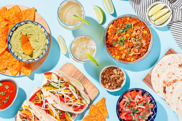 mexican food table with traditional dishes. chili con carne, tacos, tomato salsa, corn chips with guacamole. mexican feast in hard light on blue color background, top view - mexican dish imagens e fotografias de stock