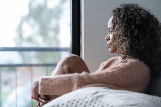 mulher negra sentada na sala de sua casa olhando para longe muito abalada por seus problemas - women depression window sadness - fotografias e filmes do acervo