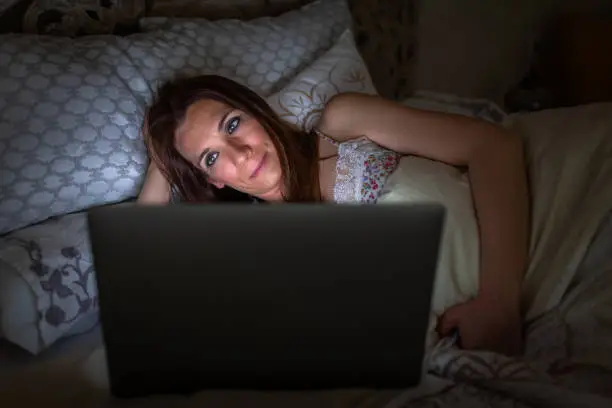 Woman in bed illuminated by the light of a switched-on laptop computer
