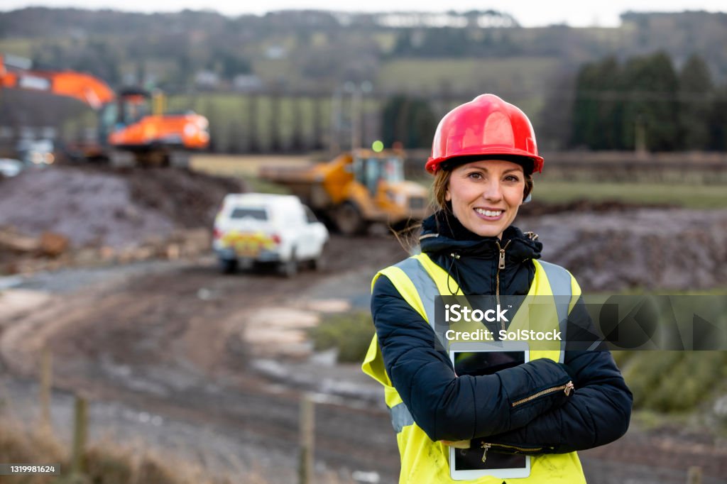 This is Where I Work A portrait of a woman, part of a survey crew looking at the camera and smiling with her arms crossed while wearing a hardhat and reflective clothing. She is working in the North East of England on a green field site and holding a digital tablet. The area where she is working is behind her. Construction Site Stock Photo