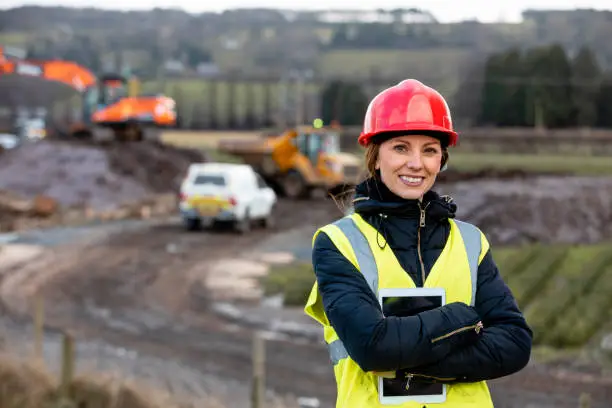 A portrait of a woman, part of a survey crew looking at the camera and smiling with her arms crossed while wearing a hardhat and reflective clothing. She is working in the North East of England on a green field site and holding a digital tablet. The area where she is working is behind her.