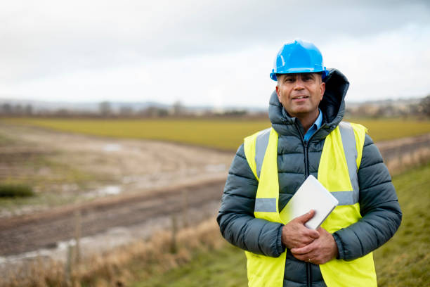 Ready to Work A portrait of a man, part of a survey crew looking at the camera and smiling while wearing a hardhat and reflective clothing. He is working in the North East of England on a green field site and holding a digital tablet. flood plain photos stock pictures, royalty-free photos & images