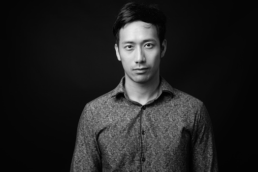 Studio shot of young handsome Asian businessman against black background in black and white