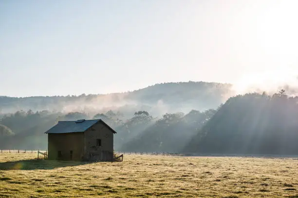 Photo of Sunrise morning silhouette of agricultural abandoned brick storage barn shed in autumn landscape farm field in rural countryside of West Virginia with fog mist sun sunrays by rolling hills