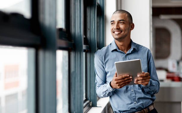 shot of a young businessman using a digital tablet while standing at a window in an office - working man imagens e fotografias de stock