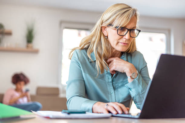 istock Serious mature woman using laptop while sitting at kitchen table 1319992770
