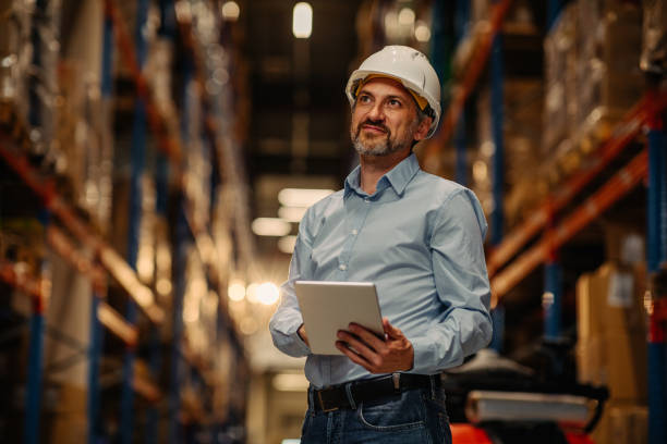 Man working in a warehouse via digital tablet Manager with safety helmet holding digital tablet in warehouse warehouse stock pictures, royalty-free photos & images
