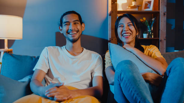 ロマンチックなアジアのカップルの男女の笑顔と笑いは、自宅で一緒にテレビでコメディ映画を見て夜のリビングルームのソファに横たわっています。 - sofa couple relaxation comfortable ストックフォトと画像