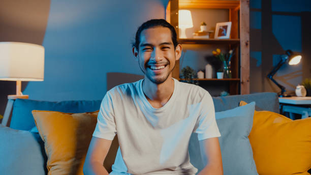 jeune homme asiatique indépendant heureux regardant le sourire d’appareil-photo et la détente gaie sur l’appel vidéo en ligne la nuit dans le salon à la maison. - men laughing portrait people photos et images de collection