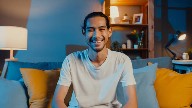 feliz jovem asiático autônomo olhando para a câmera sorrindo e alegre relaxando em chamada de vídeo on-line à noite na sala de estar em casa. - selfie - fotografias e filmes do acervo