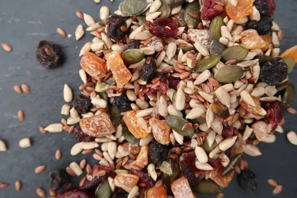 Scattered pile of edible seeds and chopped dried fruit (linseeds, pumpkin, sunflower, apricots, cranberries and blueberries). On a dark blue grey slate textured background. Healthy vegetarian lifestyle food. Flat lay from above.