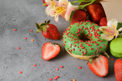 green  macaroons,1  donut,  fresh red strawberries ,  alstrameria flowers, paper  bag on a gray background