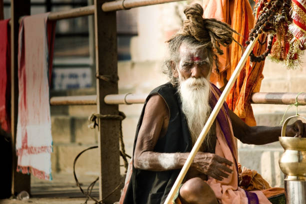 Portrait of a hindu white bearded sadhu, pilgrim or Aghori baba with a lota, braided necklace and dreadlock against ganges river ghat during day. Varanasi, India - November 01, 2016: Portrait of a hindu white bearded sadhu, pilgrim or Aghori baba with a lota, braided necklace and dreadlock against ganges river ghat during day. lota stock pictures, royalty-free photos & images